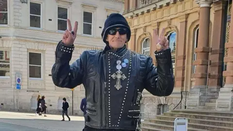 A man wearing a black leather jacket, black hat, and black glasses. He is doing a peace sign with both of his hands