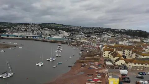 Drone shot showing a beach with boats on, wit more boats in the sea, and houses behind the beach