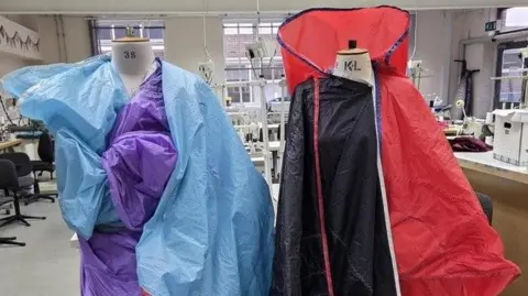 Anglia Ruskin University Two mannequins covered with large pieces of brightly coloured sail fabric, one in shades of blue and purple, the other in red, black, and white. In the background, there are rows of industrial sewing machines, chairs and tables.