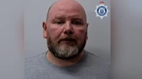 Police custody image of Kieran Cameron. He has a beard and is wearing a grey sweatshirt.