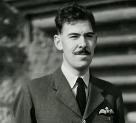 John Cruickshank A black and white picture of John Cruickshank in his RAF uniform, staring just off camera. He has a dark moustache and dark hair. He is only visible from the neck up.