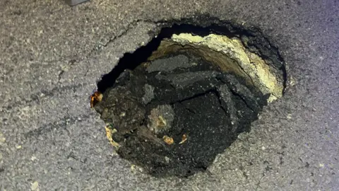 A tarmac road has partially collapsed. It is not possible to gauge the scale of the hole. 