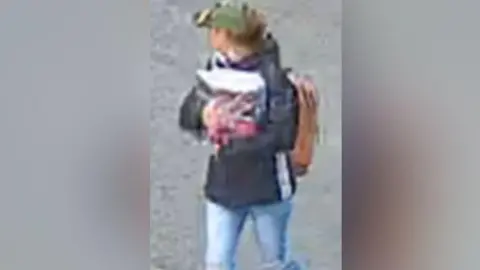 North Yorkshire Police A grainy CCTV image of a woman looking away from the camera, wearing a green cap, brown rucksack and brown jeans. She's carrying an unidentifiable object in her arms.