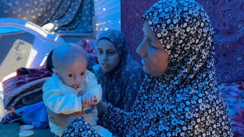 A grandmother wearing floral black and white clothing holds a tiny baby with another woman, the baby's mother, in the background, set against purple patterned cloth.