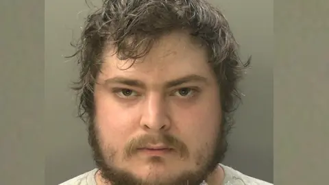 A mugshot of a man with lots of brown hair and facial hair. he is looking intensely, directly into the camera.
