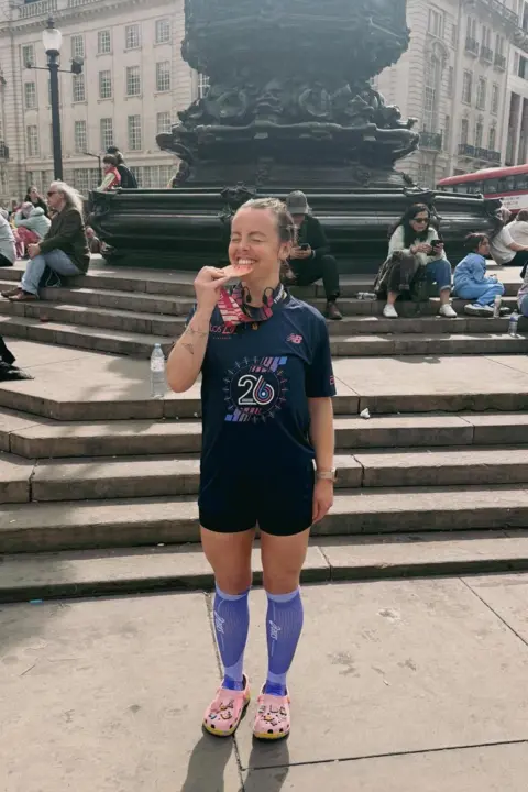 Tegan Pryce Tegan Pryce is standing in a London square in her running kit and is biting a medal she is holding and grinning.