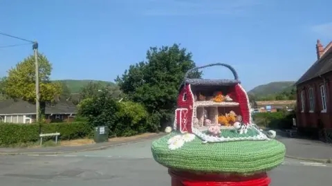 BBC Weather Watchers/George Groundhog A knitted topper on the top of a postbox next to a road, under a blue sky. The topper is the inside of a barn, with woolly farm animals.