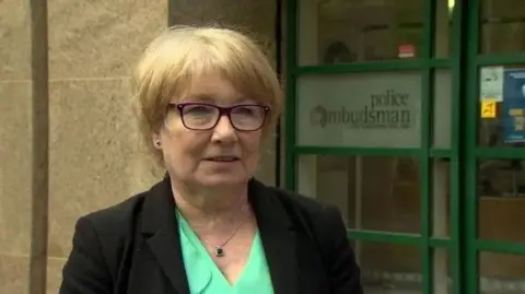 Northern Ireland's Police Ombudsman Marie Anderson in a black jacket and green top. She is wearing purple glasses and a sapphire necklace.