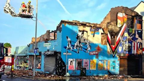 Building painted in blue with lots of murals and posters on it. It has a partly obscured road sign which says "Villiers Street" also on the left hand side. Some of the artwork on the building include multiple dogs and a large white, red and black V shape. The building has been devastated by fire and the roof has fallen in. There are firefighters on a crane in the top left of the photo. 