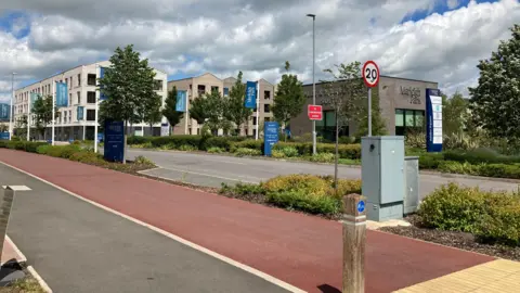 Emma Howgego/BBC A general view of a new estate. It shows three-storey buildings with flags saying "Marleigh Park" on them. In the foreground is a red cycle path and grey footpath. 