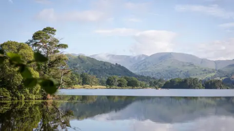 National Trust/PA Media A panoramic view of a still lake, with hills and trees reflected in its vast waters.
