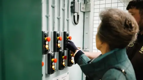 Robbie Dark Photography Princess Anne is standing by a control panel with several red buttons and prepares to press one to open the enclosure and let the gorillas in.