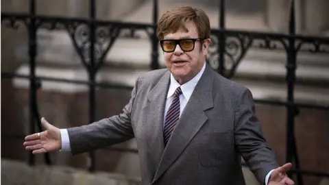 PA Media Elton John with short dark hair and black-framed glasses wearing a grey jacket, white shirt and striped tie, with his arms held out. He is standing in front of some black railings.