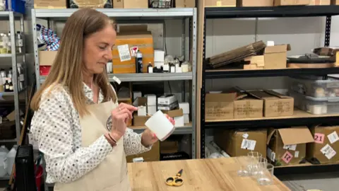 A woman in an apron and patterned blouse holding a wick of a candle in a glass jar