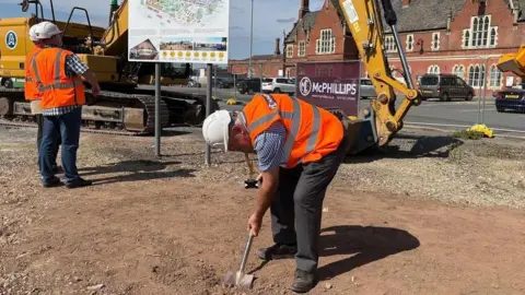 Two men wearing high-viz jackets, stood on a mound of soil - one of the men is bending over sticking a spade in the ground.