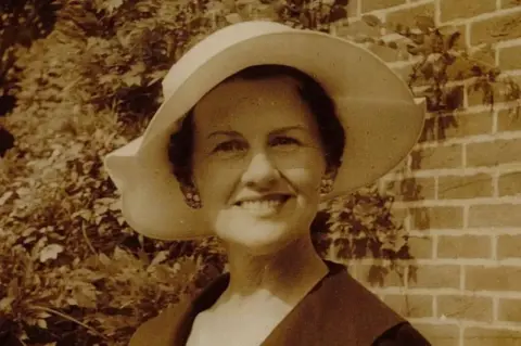 Muriel McKay photographed in a black and white sepier picture. She is wearing a cream summer hat and a dark jacket open at the neck, and cluster earrings. She is smiling at the camera and standing in front of a brick wall with a trailing plant like wisteria growing in the background.