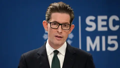 PA Media Ken McCallum, Director General of MI5 - a middle-aged man with slicked back dark brown hair, and wearing dark-rimmed rectangular glasses. He is wearing a dark suit, white shirt and navy blue tie.