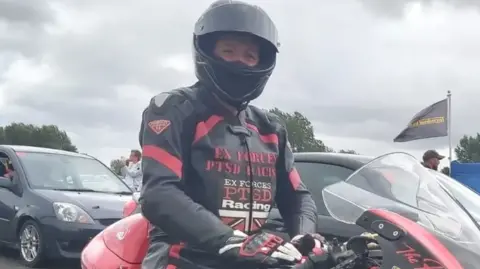 A man wearing black and red bike leathers and a helmet sits on his red motorcycle at an event, with cars in the background.