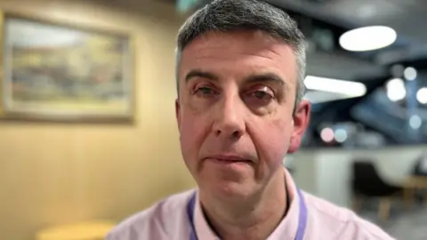 Kristian is wearing a pink shirt and is looking at the camera with a neutral expression. Behind him on the left is a beige wall with a painting, and office chairs in the background on the right of the picture.