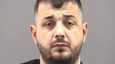 A police custody photo of Ibrahim Sollaku against a grey background. He has a round face with a neatly-trimmed full dark-brown beard and short brown hair. He is looking at the camera with a furrowed brow.