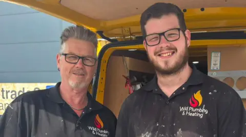Jaimielee Rendall/BBC Brandon and Tony Wild wearing black polo shirts with Wild Plumbing and Heating written on them. They are standing in front of the back of a van, which is open.