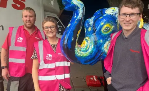St Giles Hospice Two men and one woman in pink hi-vis St Giles jackets smile as they stand next to the Vincent Van Gogh themed elephant
