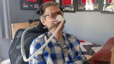 Richard is sitting in a bedroom. He has a tube over his nose. He wears glasses. There is a Manchester United pillowcase on the bed. 