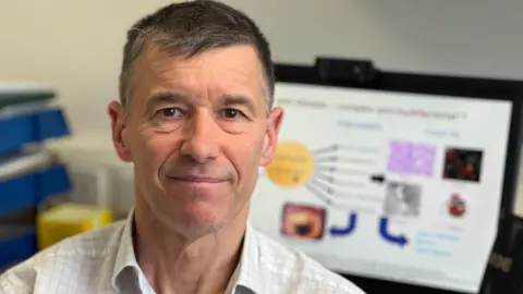 Prof John Wilding sits in front of a computer, looking at the camera. He is wearing a white checked shirt and has short dark/grey hair.