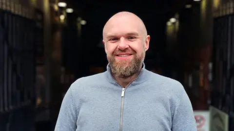 Durham Markets James Wilson is smiling at the entrance of Market Hall in Durham. He has long beard and a grey zip-up top.