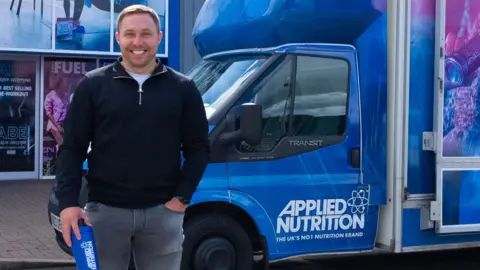 Tom Ryder, who is smiling at the camera, is standing in front of van with Applied Nutrition branding and holding an Applied Nutrition branded water bottle.