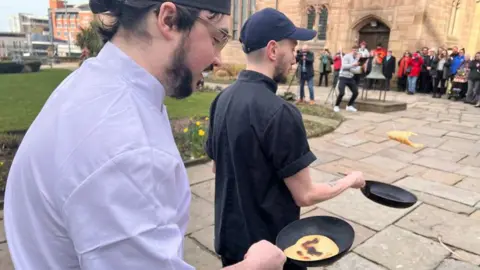 Two chefs are tossing pancakes in black frying pans as the run past Liverpool Parish Church