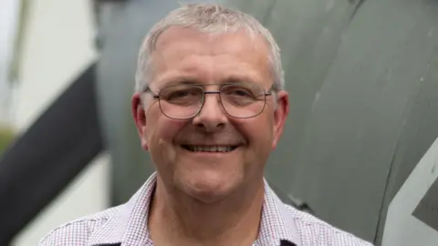 David Price David Price standing in front of the Spitfire he built. He has short white hair and glasses and is wearing a shirt with a chequered pattern. Behind him is the spitfire is painted a brown-grey colour.