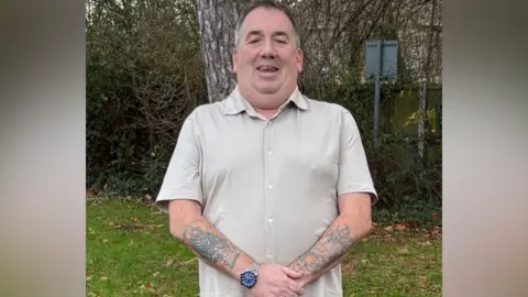 A man wearing a beige shirt is standing in a garden. He has tattoos all over his arms and is wearing a watch.