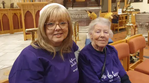 BBC Kathleen and Sharon smiling at the camera. Both are wearing purple t-shirts. Sharon has blonde hair and is wearing glasses.