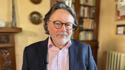 A middle-aged man with grey swept-back hair and round black glasses smiles slightly at the camera. He is wearing a suit and a pink open-neck shirt.