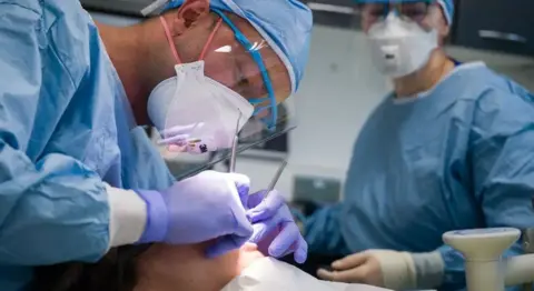 Stock image of dentists in protective gear wearing gloves and delicately inserting metal items to someone's mouth and teeth