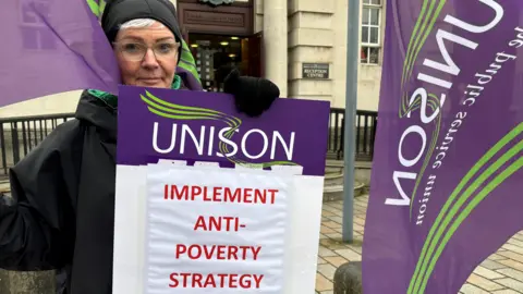 A woman holds a placard which says 'Implement anti-poverty strategy' in red text. Above this it says 'Unison' in white text on a purple background. The Royal Courts of Justice are behind her