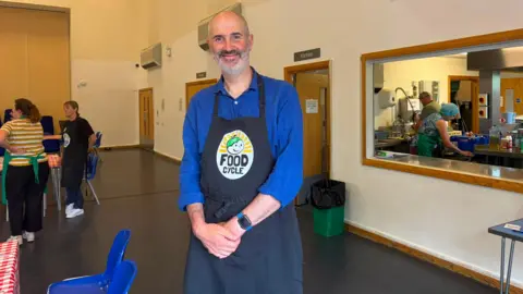 A tall man with a grey beard wearing a dark blue shirt and a black apron reading 'FoodCycle' is smiling whilst standing in a community hall in front of a kitchen area.