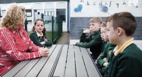 St Michaels RC Primary School Head teacher Joanna Taylor who has blonde and glasses and is wearing a red top. She is sat at a picnic bench and has her head turned away from the camera as she talks to a young girl in green and yellow school uniform. At the other side of the bench, four boys in the same uniform are sat looking at the pair.