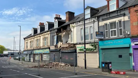 A row of shops along the side of a road. The front of the first floor of one of the buildings has collapsed onto the pavement and road below. Wire fencing has been put up to cordon off the area.
