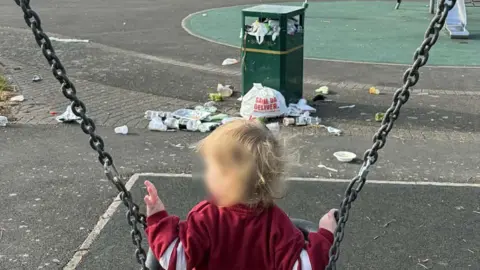 A child sat on a swing in a park. Infront of the child is an overflowing public bin and litter on the floor.