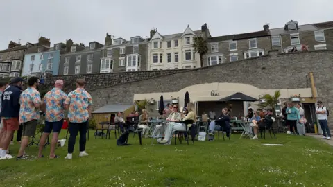 Ellie Pizey The restaurant on a grey evening. There are three men in flowery shirts standing on the grass in the foreground. Many people are sitting at tables with drinks. The building is built into a wall and above it is a row of two and three-storey houses. 