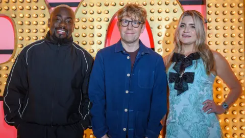 Comics Andrew Mensah, Josh Widdicombe and Harriet Kemsley seen on Live at the Apollo earlier this year, standing on stage with the programme logo in lights in the background