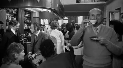 Getty Images A black and white archive image taken inside a pub full of people chatting in groups. On the right, a man with thinning hair smiles as he holds a teacup to his lips with the sauce in his other hand. He is wearing a jumper over a shirt and tie. The elaborate, three-sided bar, to the left, has a wooden ceiling. Two middle-aged men sit to one side, while in the centre of the room a group of women are in conversation.