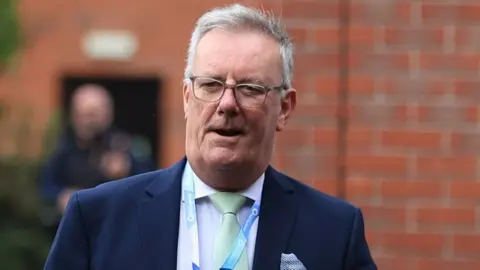 Mike Nesbitt has grey hair and glasses and is wearing a dark blue suit jacket, light shirt and light blue tie