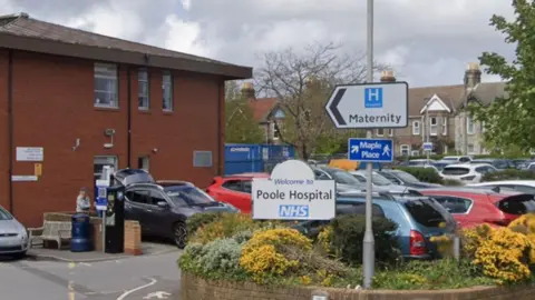 Google Google Street View of St Mary's hospital in Poole. The two storey red brick building is surrounded by a car park full of cars. At the entrance are two signs which say Welcome to Poole Hospital and Maternity.