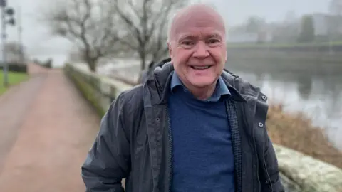 BBC A grey haired, balding man with a dark jacket and blue jumper and shirt stands in front of the River Nith in Dumfries