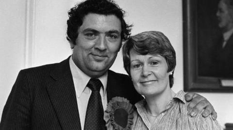 John Hume stands to the left with his arm around his wife Pat. The image is black and white. He has dark hair and a dark suit with a large political-style rosette attached to his blazer. Pat has short dark hair and is wearing a patterned shirt.