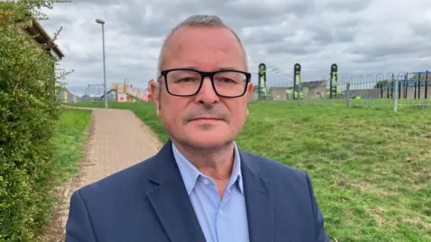 A head-and-shoulders shot of Lee Barron, wearing a light-blue shirt and dark-blue jacket. He is standing on a path in front of a playground. He has glasses and short grey hair and is looking at the camera.