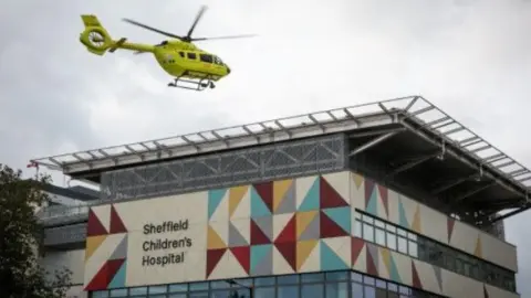 Sheffield Children's Hospital A building with glass windows and a sign that reads 'Sheffield Children’s Hospital' in black letters on a multi-coloured panel. Above the building, a yellow helicopter is flying.